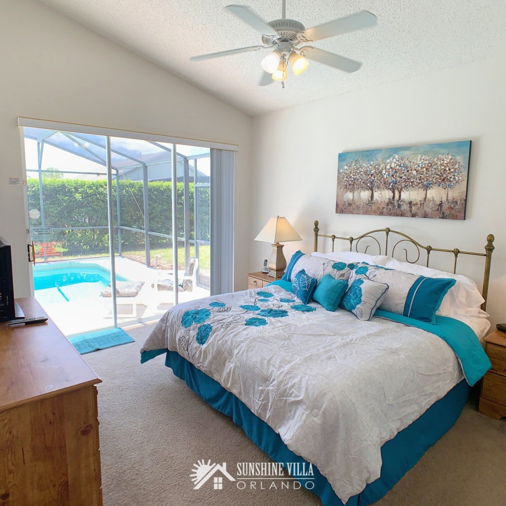 Master Bedroom at Sunshine Villa at Glenbrook Resort, a short-term vacation rental home in Orlando near Walt Disney World. A king sized bed is luxuriously filled with pillows and a large fluffy comforter. An extra wide glass sliding door looks from the room to a private in ground pool.